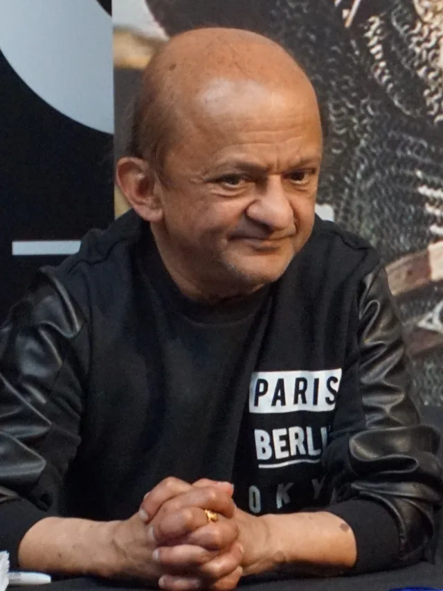 Head and shoulders photograph of Kiran Shah, a bald man in a black top with leather sleeves, seated with his hands clasped and a slight smile.