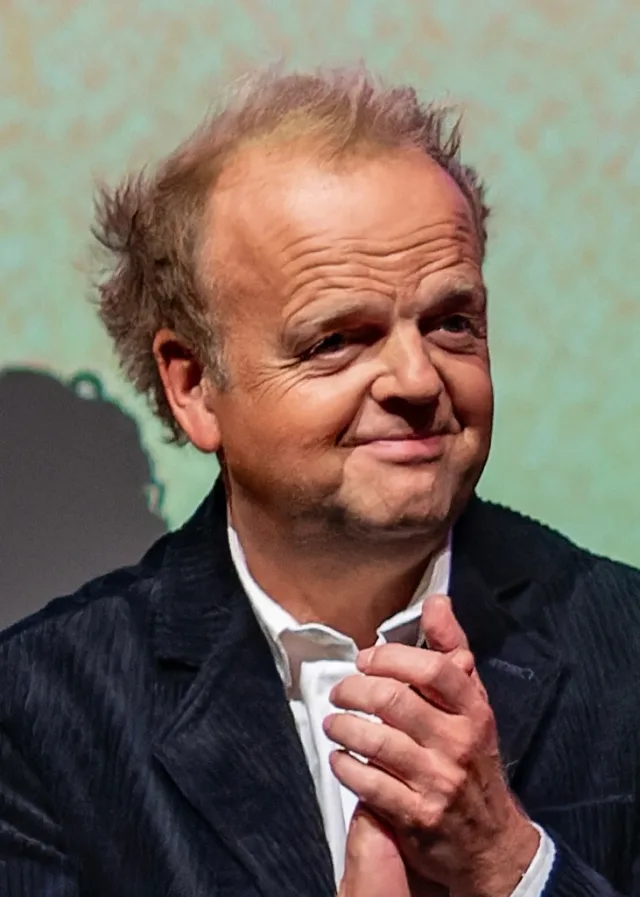 Man with tousled thinning hair and a wry smile, in a dark corduroy jacket and white shirt, hands pressed together.