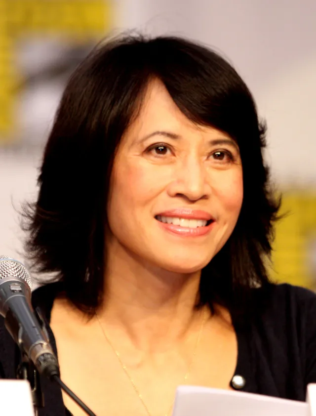 A smiling woman with shoulder-length dark hair and a fringe, in a dark cardigan, seated at a microphone at a convention panel.