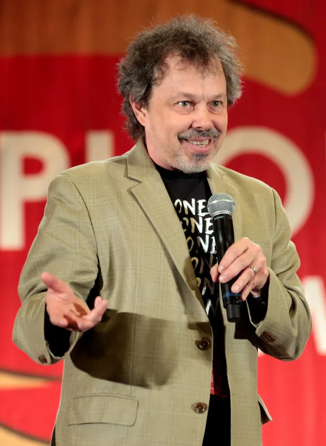 Curtis Armstrong smiling and gesturing as he speaks into a microphone, in a tan jacket over a black t-shirt.