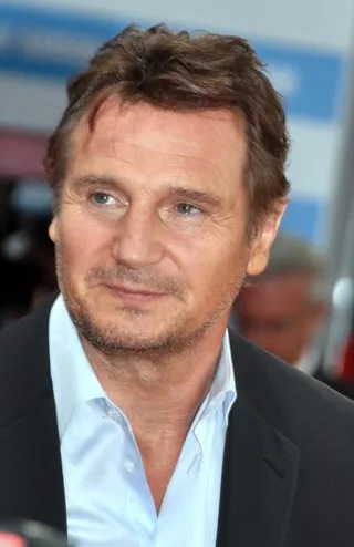 Close-up photograph of Liam Neeson at a festival, with short brown hair and stubble, wearing a pale blue open-collared shirt under a dark jacket.
