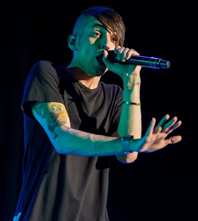 Singer in a black t-shirt performing on a dark stage, singing into a microphone with one hand outstretched