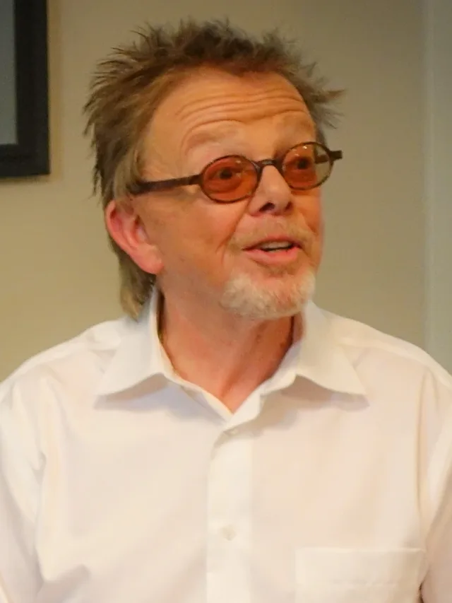 Head and shoulders photograph of a man with spiky hair, a grey goatee and round tinted glasses, wearing a white shirt and speaking.