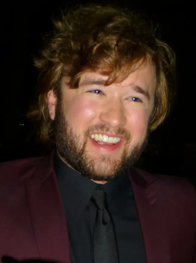 Haley Joel Osment laughing, with tousled hair and a beard, in a maroon jacket and black shirt.