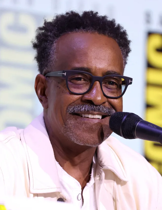 A smiling man with short curly hair, dark-framed glasses and a grey moustache, speaking into a microphone in a cream jacket.