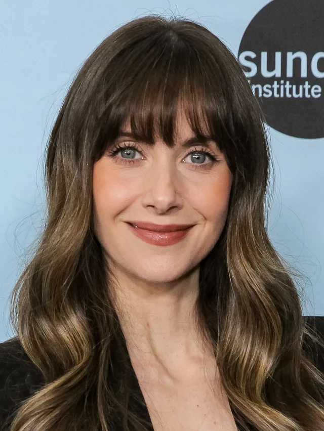 A woman with long brown hair and fringe, with blue eyes and a faint smile, in front of a pale blue festival backdrop.