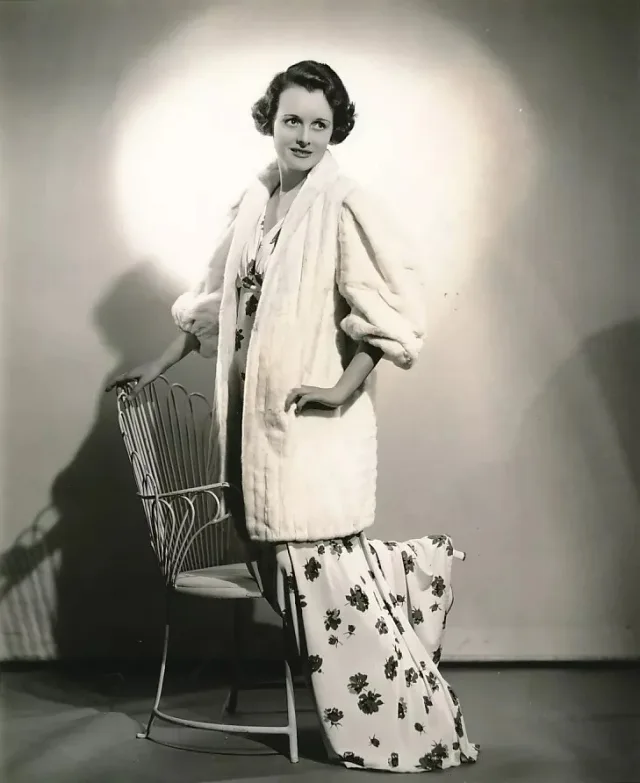 Black and white studio photograph of a dark-haired woman in a white fur jacket and floral gown, standing beside a wire chair in a spotlight.