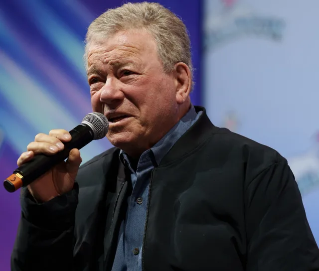 William Shatner speaking into a handheld microphone, wearing a dark zip jacket over a blue shirt.