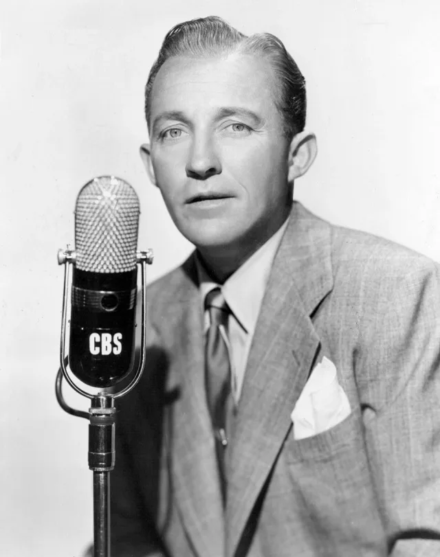 Black and white studio portrait of a man in a light checked jacket and tie, seated beside a vintage CBS radio microphone.