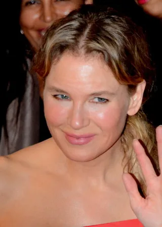 Close photograph of a woman with swept-back light brown hair, smiling slightly and raising a hand, wearing a red strapless top at a public event.