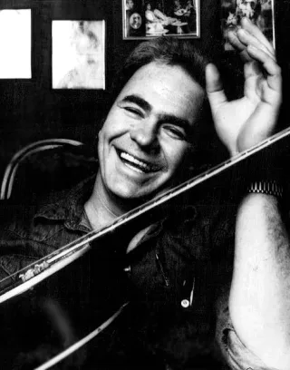 Black and white photograph of a smiling man with dark hair holding an acoustic guitar across his body and raising one hand