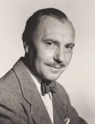 Black and white studio portrait of a man with a moustache and receding hair, wearing a tweed jacket and a spotted bow tie.