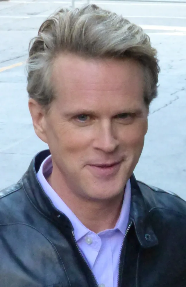 Close portrait of Cary Elwes outdoors, with swept-back grey-blond hair, a lilac collar and a dark leather jacket.