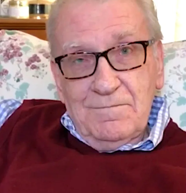Close photograph of an elderly man with white hair and tortoiseshell glasses, in a burgundy jumper over a blue checked shirt, seated on a floral sofa.