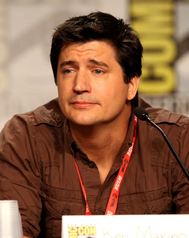 Ken Marino with dark hair and a brown shirt, seated at a convention panel table with a microphone, looking slightly pensive