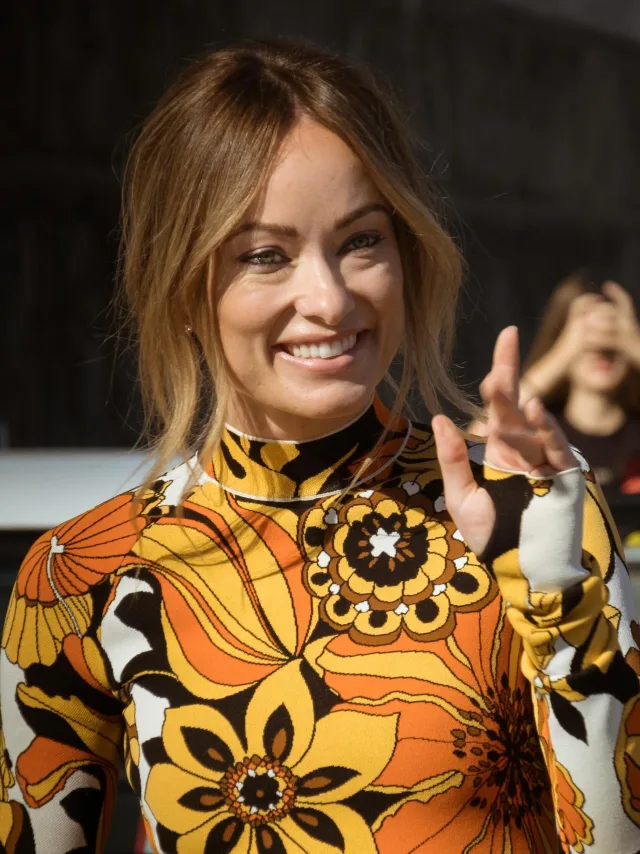 Olivia Wilde smiling and waving in a bold orange, yellow and black floral top, photographed outdoors.