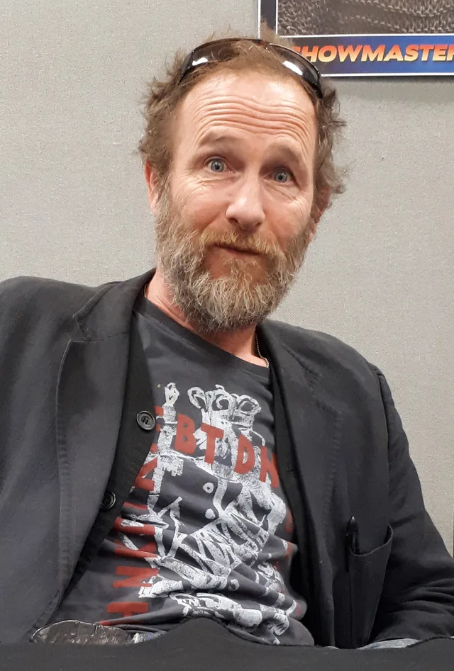 Bearded man with tousled hair and sunglasses pushed up on his head, wearing a dark jacket over a graphic t-shirt, seated at a convention table.
