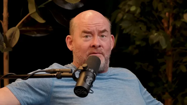 A bald man with a reddish beard and a raised eyebrow, in a light blue t-shirt, seated behind a studio microphone with plants in the dark background.