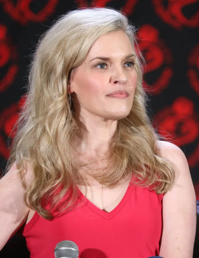Portrait of Kari Wahlgren with long wavy blonde hair, in a red top, seated at a panel with a microphone in front of her.