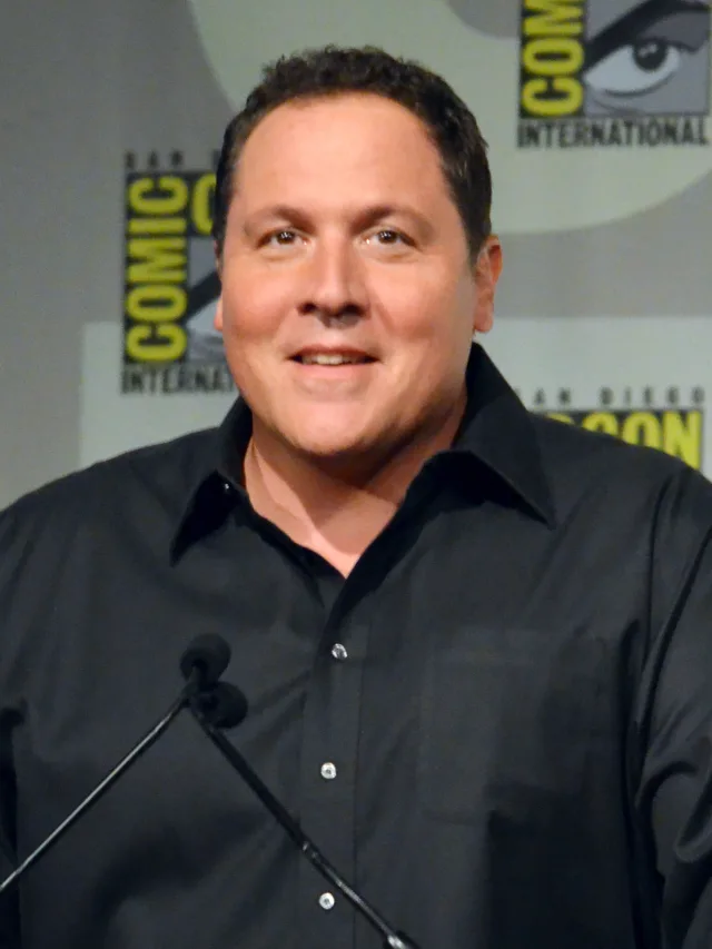 Head and shoulders photograph of a smiling man with short dark hair in a black collared shirt, speaking at a microphone at a convention panel.