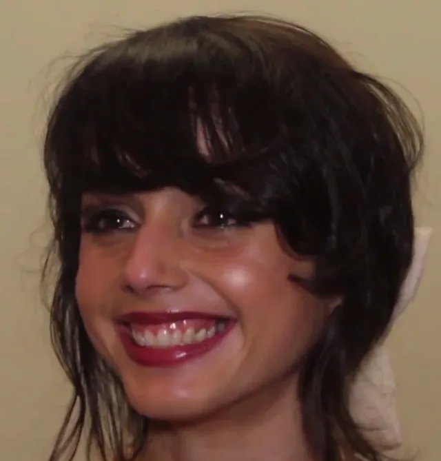 Close photograph of a smiling woman with dark fringed hair and red lipstick against a beige wall.