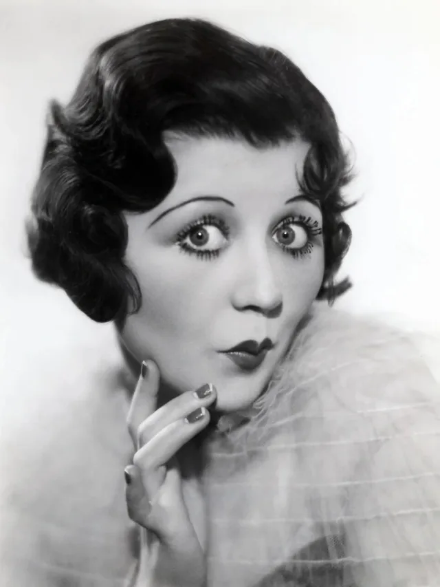 Black and white studio portrait of a young woman with short wavy dark hair and wide eyes, lips pursed, one hand raised to her chin, in a soft feathered wrap.