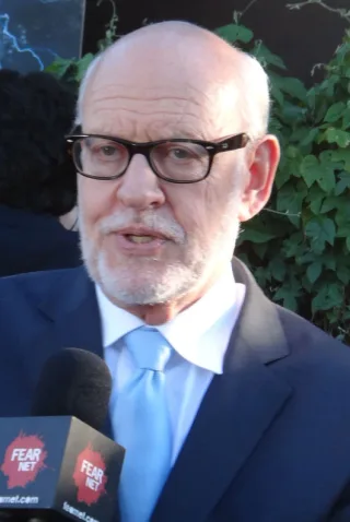 Close photograph of an older man with a white beard and dark-framed glasses, in a navy suit and pale blue tie, speaking to a microphone.