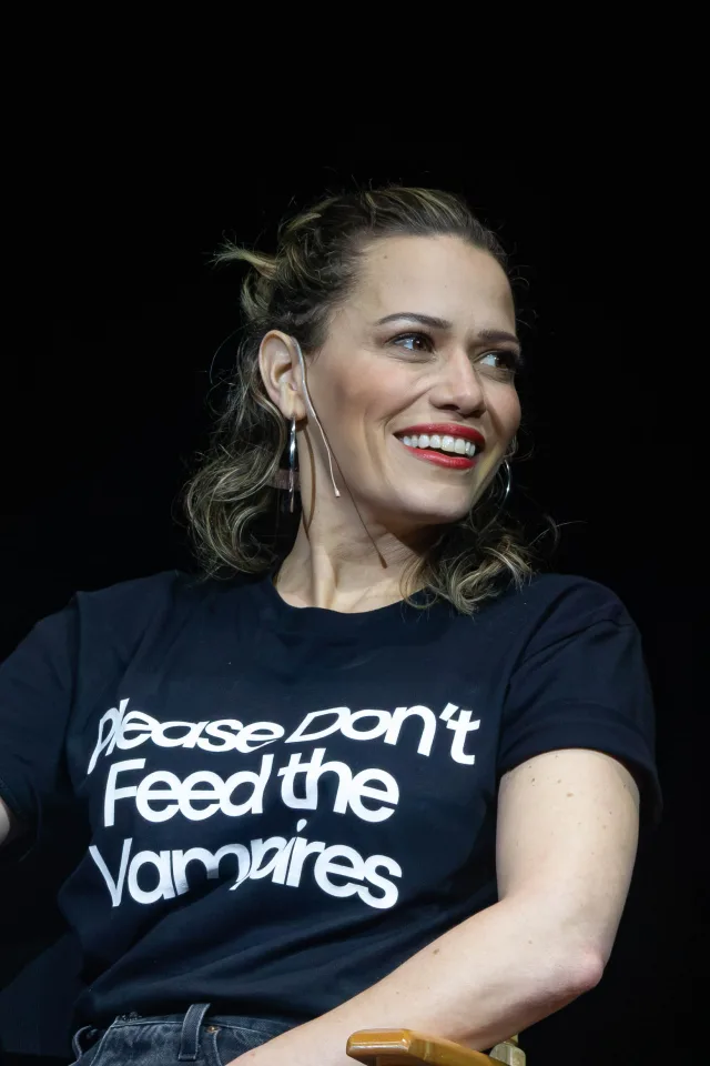 Bethany Joy Lenz smiling and glancing to one side on a dark stage, in a black t-shirt with white lettering.