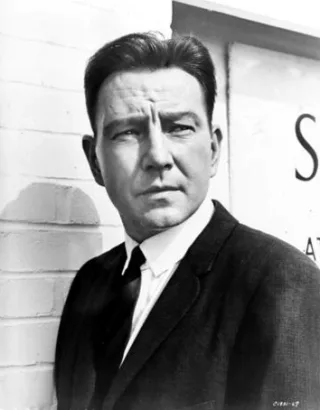 Black and white publicity portrait of a man in a dark jacket and tie, standing against a brick wall with a serious, sideways look.