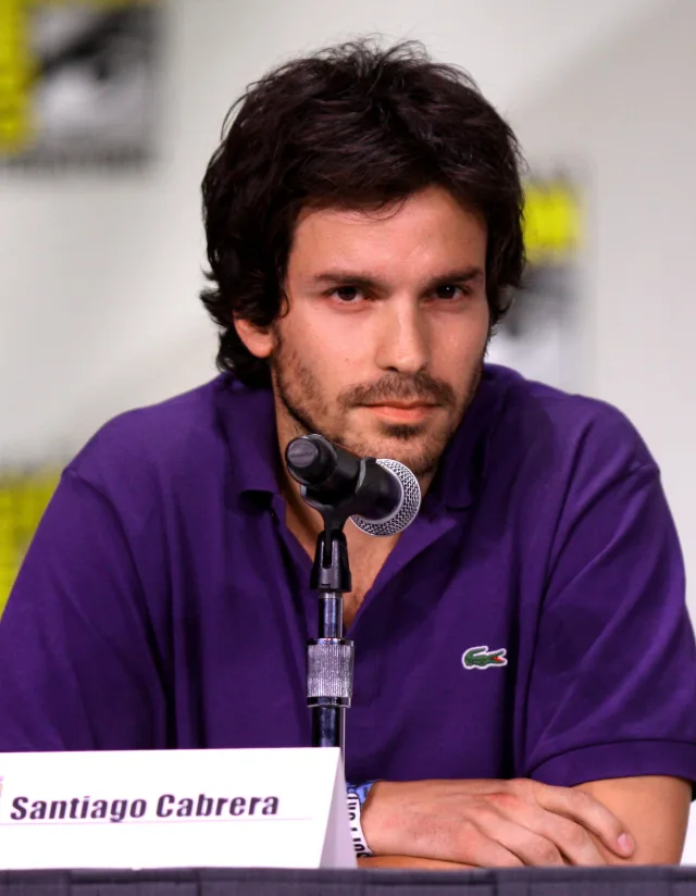 Santiago Cabrera with dark tousled hair and stubble, in a purple polo shirt, seated at a panel table behind a microphone.