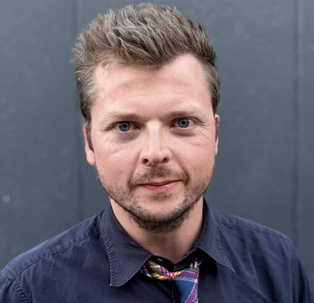 Head and shoulders photograph of a man with short spiky light brown hair and stubble, wearing a navy shirt and a patterned tie, looking straight at the camera.