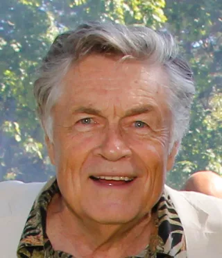 Close photograph of Art Hindle smiling outdoors, with swept-back grey hair and a patterned shirt under a cream jacket.