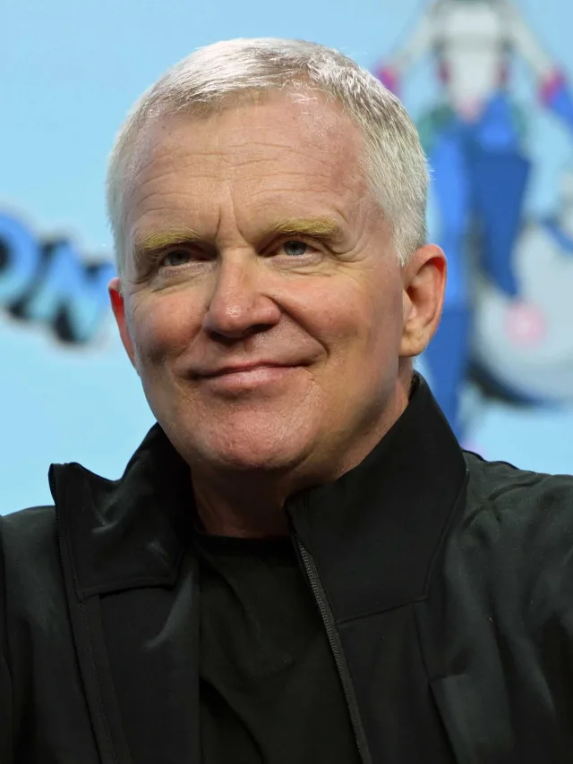 Close-up photograph of Anthony Michael Hall, a man with short white hair, smiling slightly, in a black zip jacket against a pale blue backdrop.