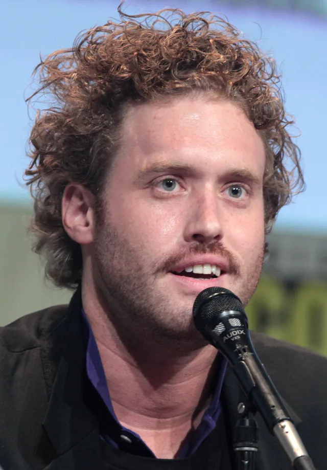 Portrait photograph of a man with curly auburn hair and light stubble speaking into a microphone, wearing a dark jacket over a purple shirt at a convention panel.