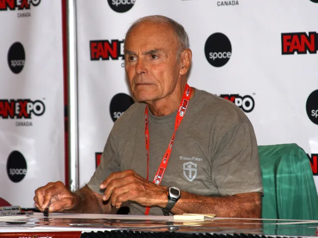 John Saxon seated at a signing table in a grey t-shirt and red lanyard, in front of a convention backdrop.