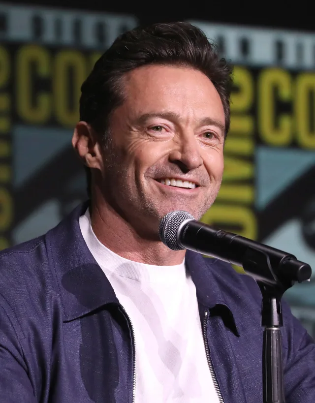 Hugh Jackman smiling at a convention panel microphone, in a navy zip jacket over a white t-shirt.