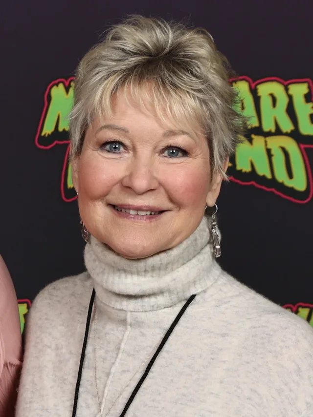 Head and shoulders photograph of Dee Wallace, smiling, with short blonde hair and a cream polo-neck jumper, in front of a dark event backdrop.