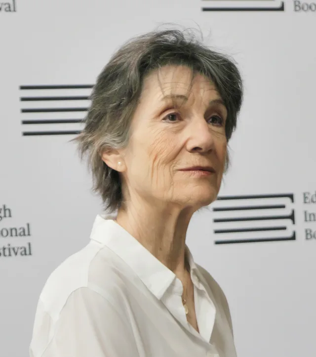 Harriet Walter in a white shirt, looking to the side in front of a book festival backdrop, with short grey hair.