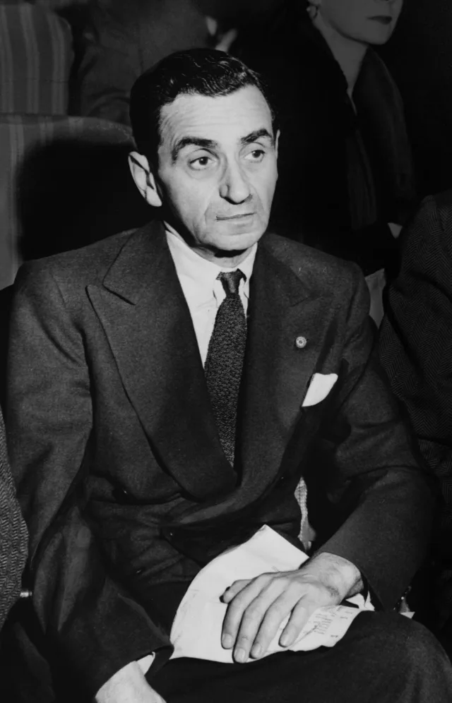 Black and white press photograph of a dark-haired man in a double-breasted suit and knitted tie, seated in an audience with papers in his lap.