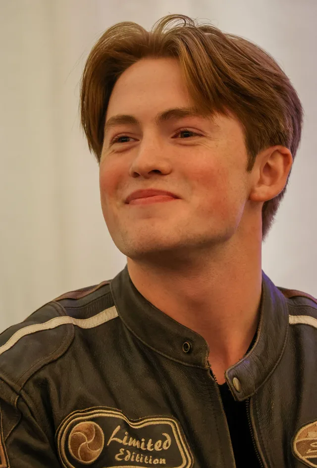 Young man with swept auburn hair, smiling and glancing to one side, wearing a brown leather jacket at a panel event.