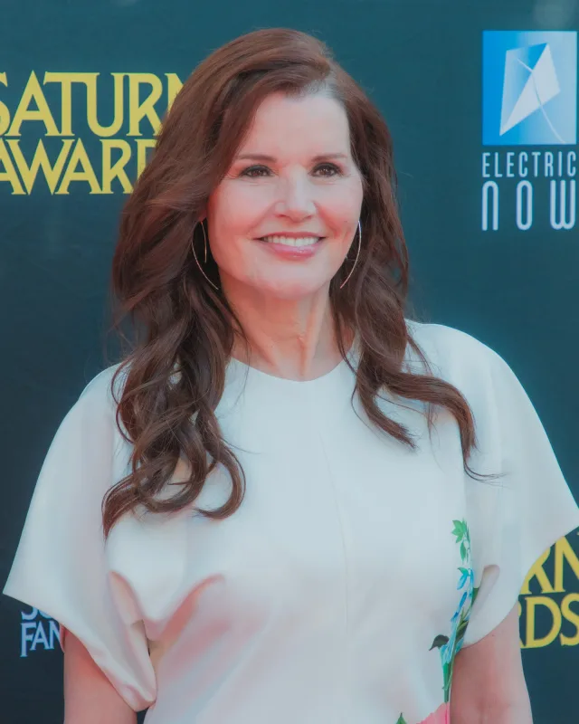 A smiling woman with long wavy auburn hair and hoop earrings, in a white short-sleeved top, at an awards event backdrop.