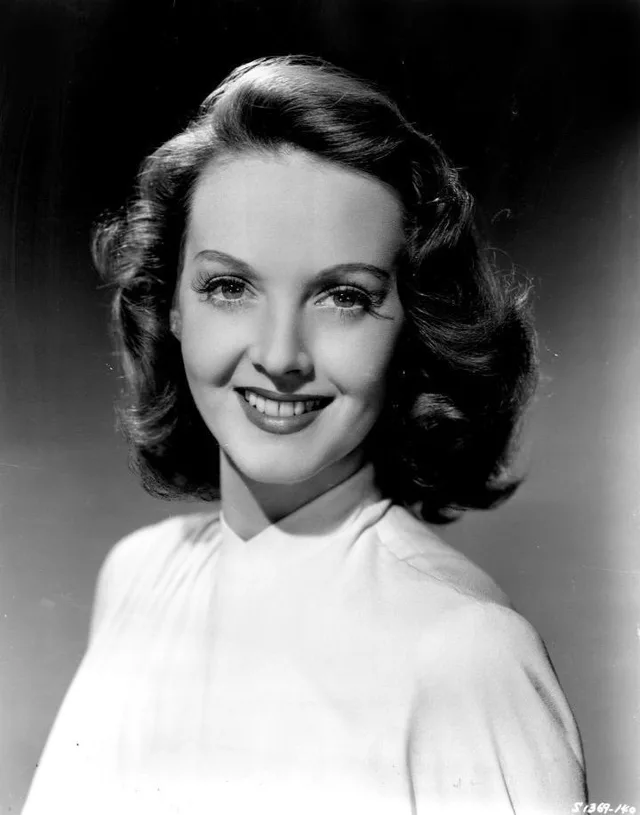 Black and white studio portrait of a smiling young woman with wavy shoulder-length hair, wearing a pale high-necked top.