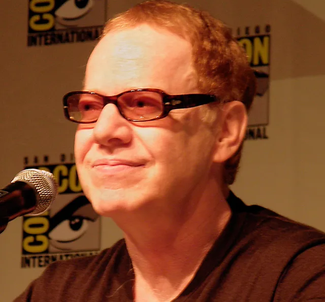 Danny Elfman speaking at a convention panel, wearing dark-framed tinted glasses and a dark top, with a microphone in front of him.