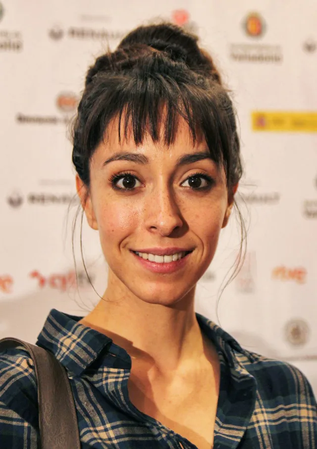 Portrait of a smiling woman with dark hair in a bun and fringe, wearing a blue checked shirt, in front of a sponsor backdrop.