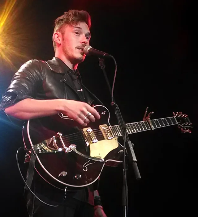 Sam Palladio singing into a microphone on stage while playing an electric guitar, wearing a black leather jacket.