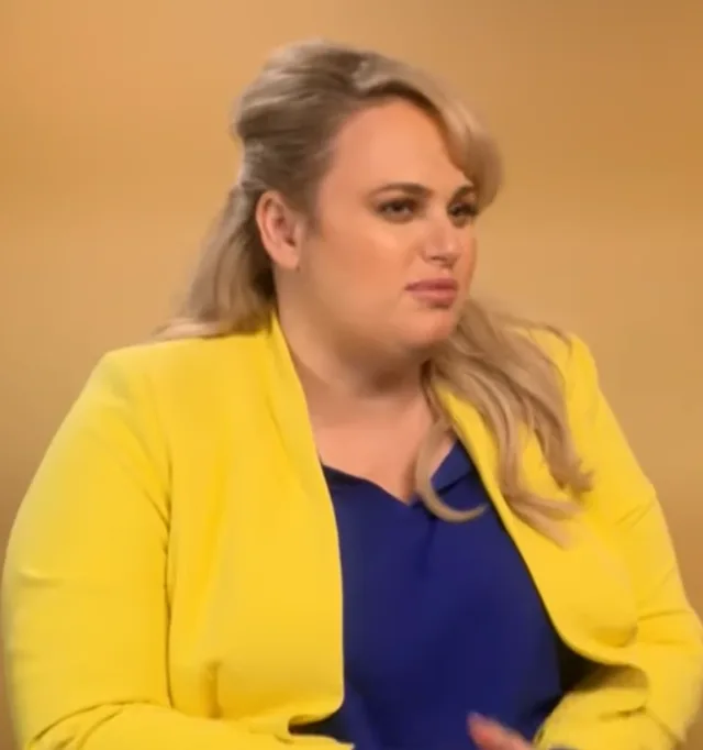 Rebel Wilson in an interview setting, with blonde hair swept back, wearing a yellow blazer over a blue top.