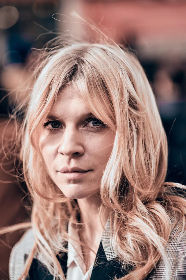 Close-up portrait of a woman with wavy, strawberry-blonde hair and a fringe, wearing a checked jacket over a white collar, looking at the camera.