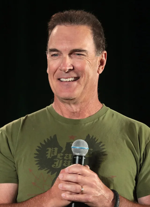 A smiling middle-aged man with short brown hair, wearing an olive green t-shirt and holding a microphone in both hands against a dark background.