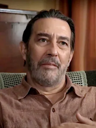 Middle-aged man with swept-back dark hair and a grey beard, in a rust-coloured shirt, seated and looking thoughtfully off to one side.