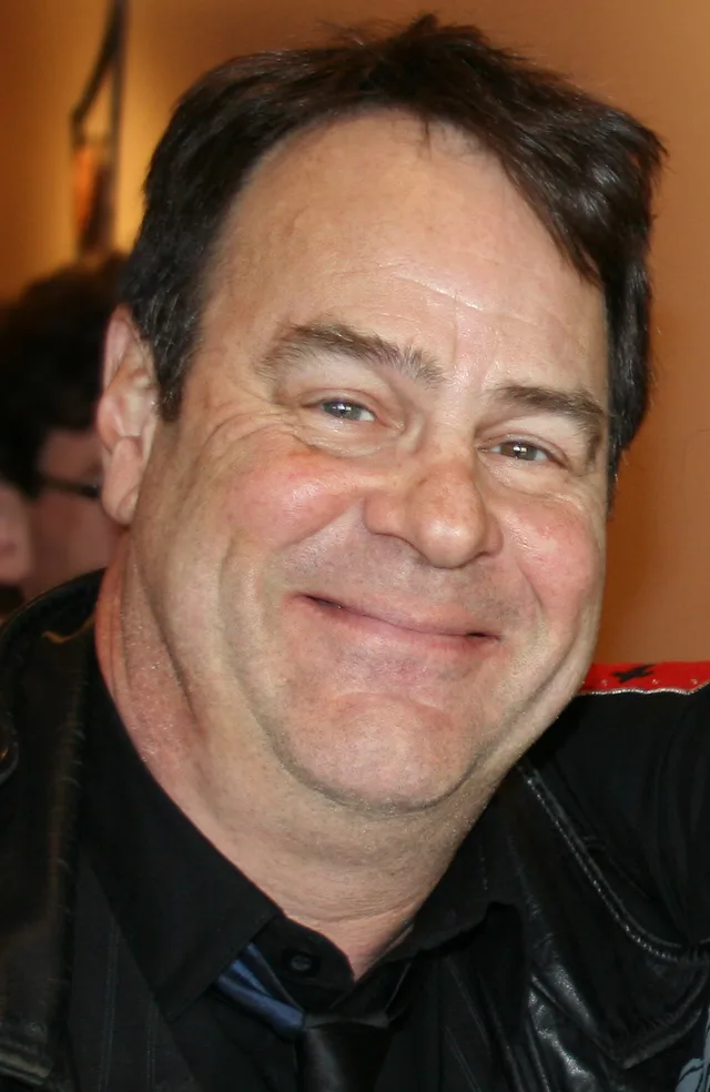 Close colour photograph of a smiling man with dark swept hair, wearing a black shirt and a black leather jacket against a warm orange background.